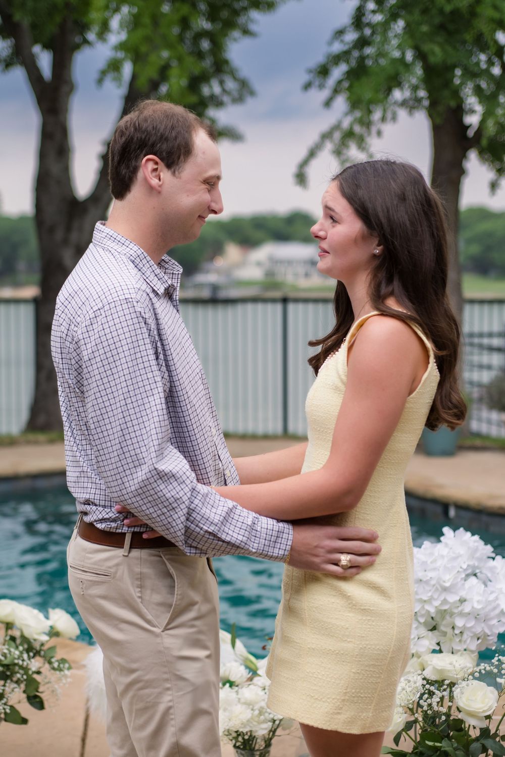 Two people share an intimate moment outdoors by a pool with trees and white flowers in the background.