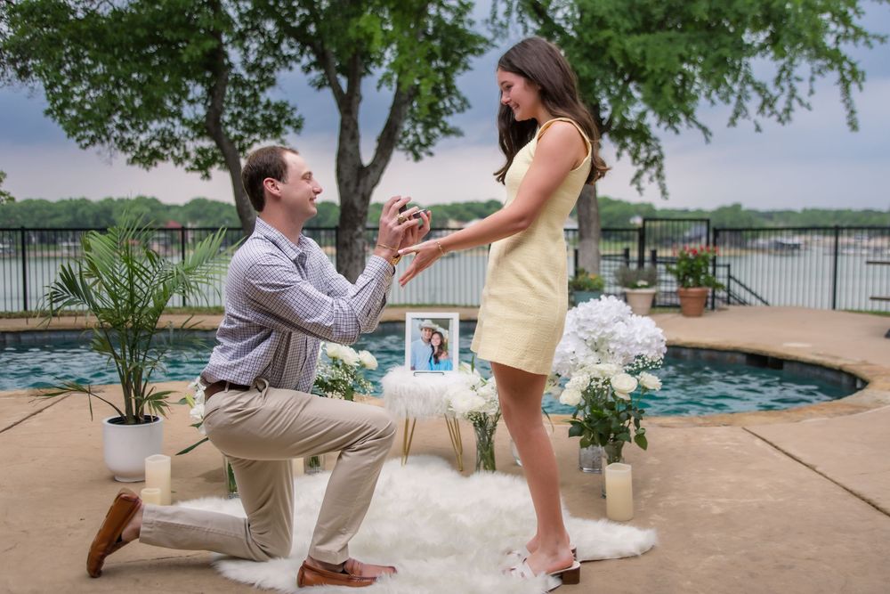 A romantic proposal scene by a poolside with trees and water in the background on a sunny day.