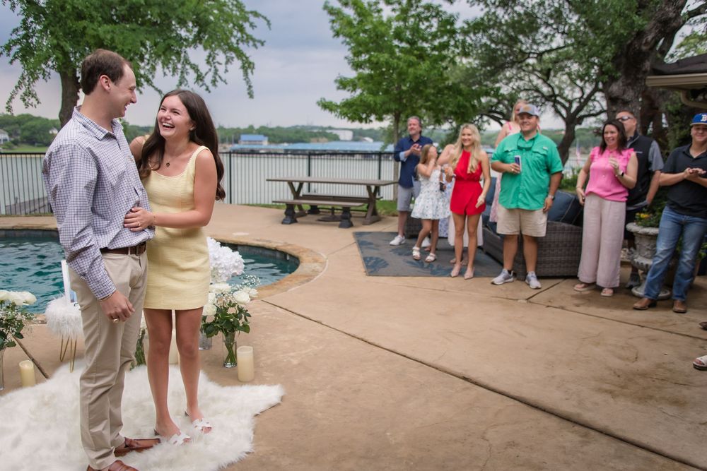 Two people stand speaking closely on a patio near a pool while a small group watches nearby at an outdoor event.