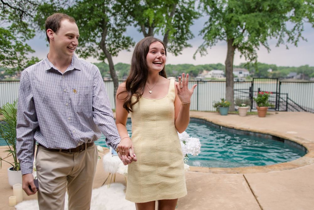 A couple stands holding hands and laughing together by a poolside with a lake view in the background.