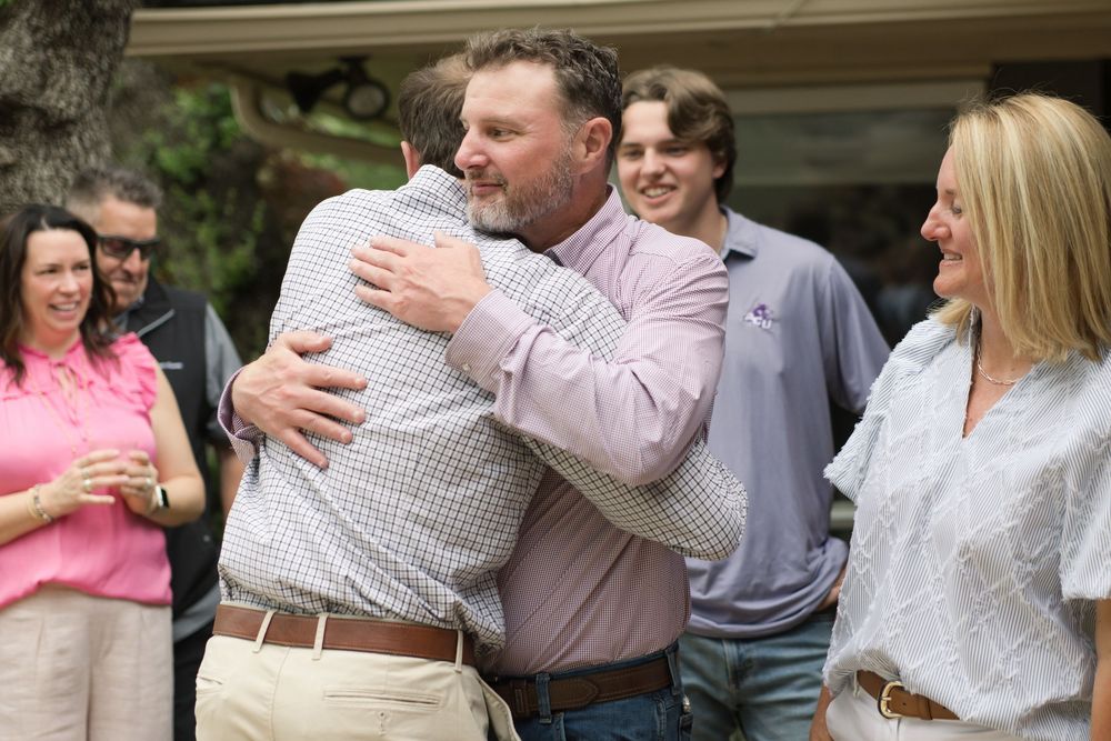 A heartfelt embrace is shared between two people at an outdoor gathering as others look on with smiles.