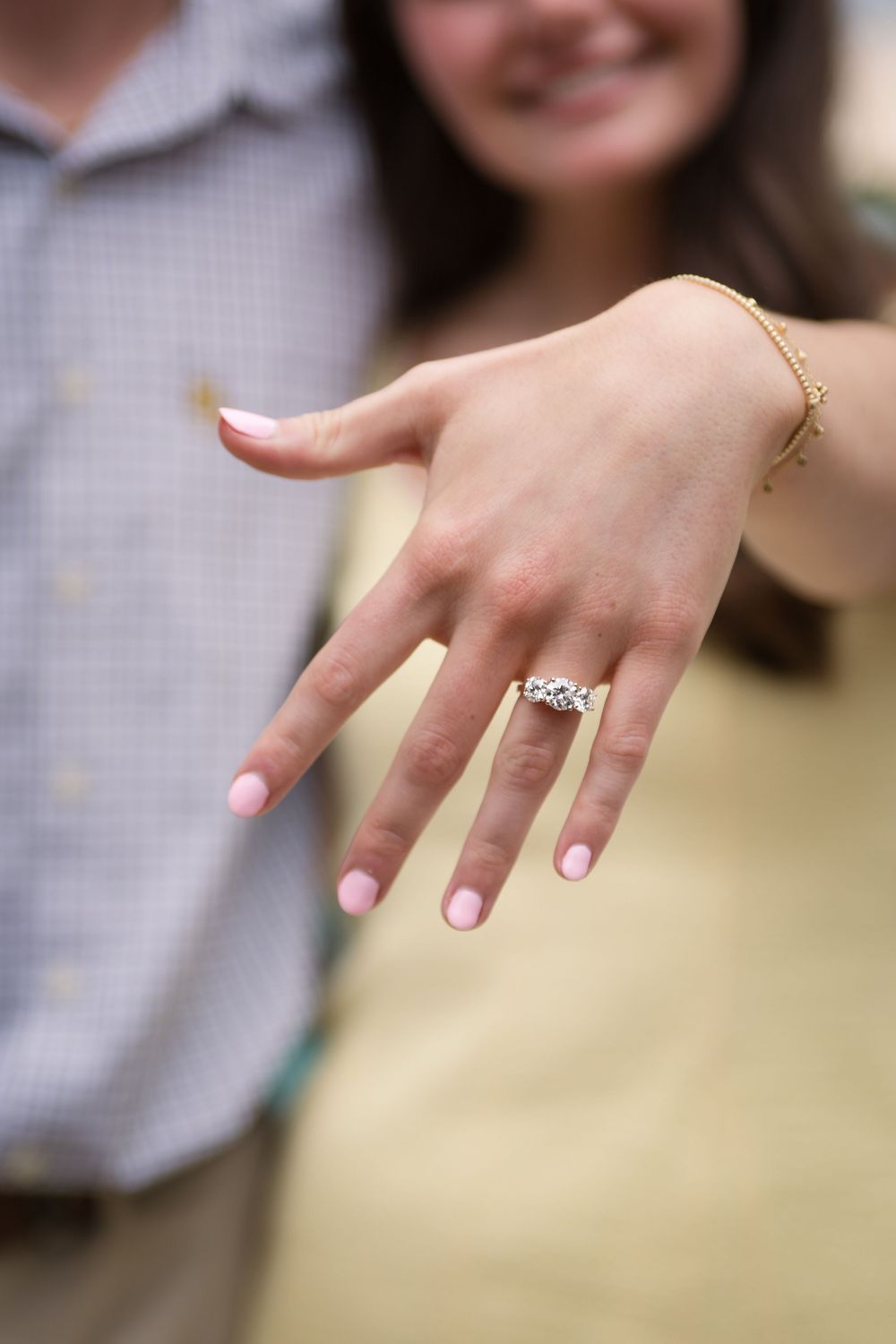 Hand displaying an elegant diamond engagement ring with a delicate gold bracelet and light pink manicure.
