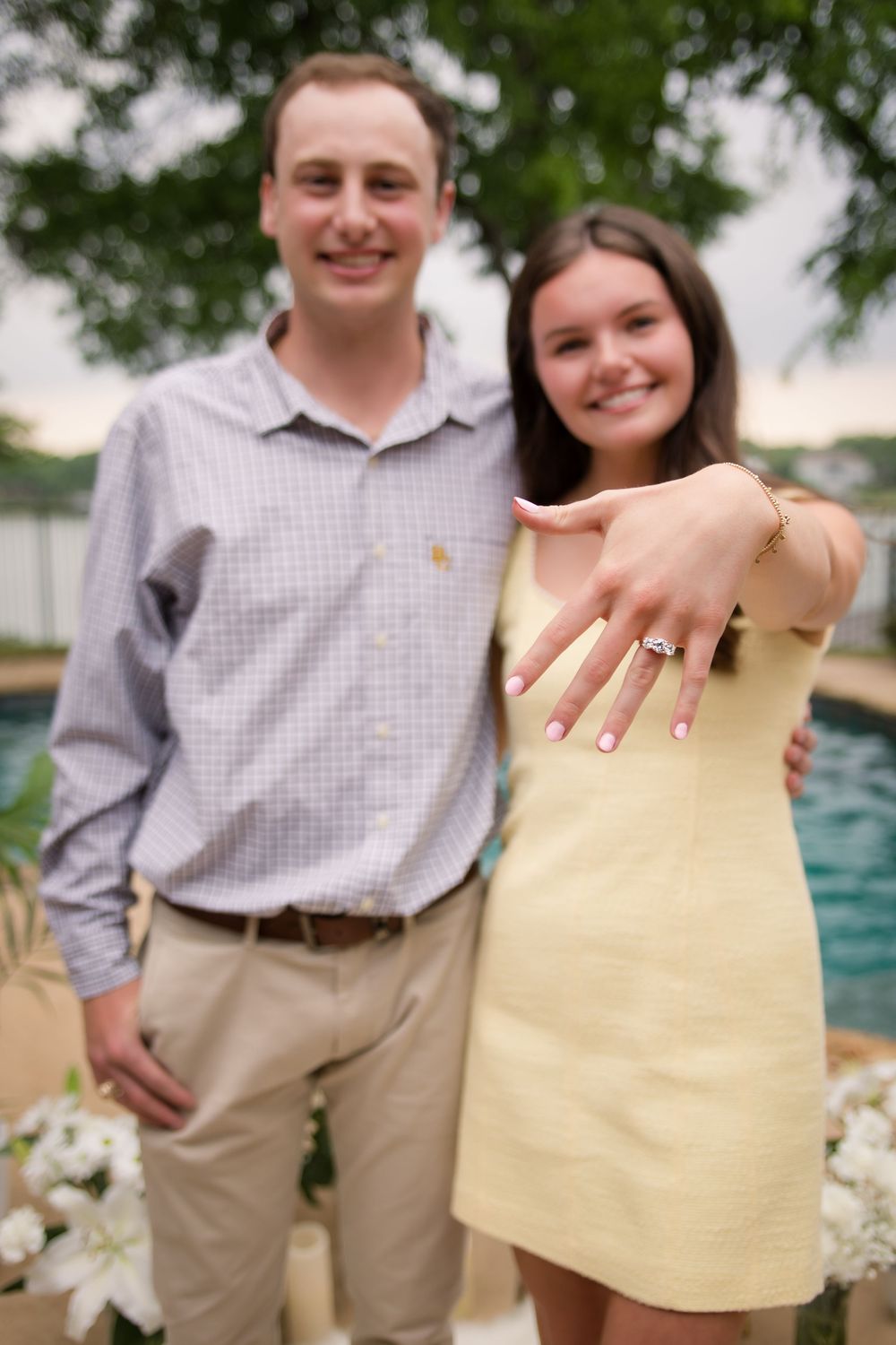 A newly engaged couple stands near a pool with a swimming pool and greenery in the background.