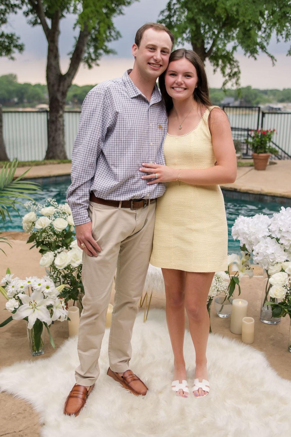 A couple poses outdoors by a waterfront on a decorated patio with white flowers and candles.