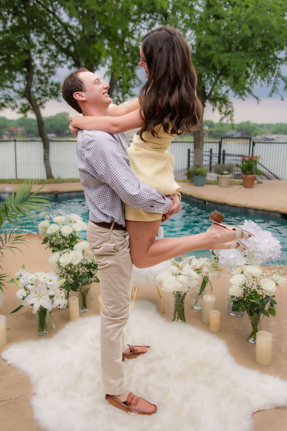 A romantic engagement scene featuring a joyful lift by a pool surrounded by white floral arrangements.