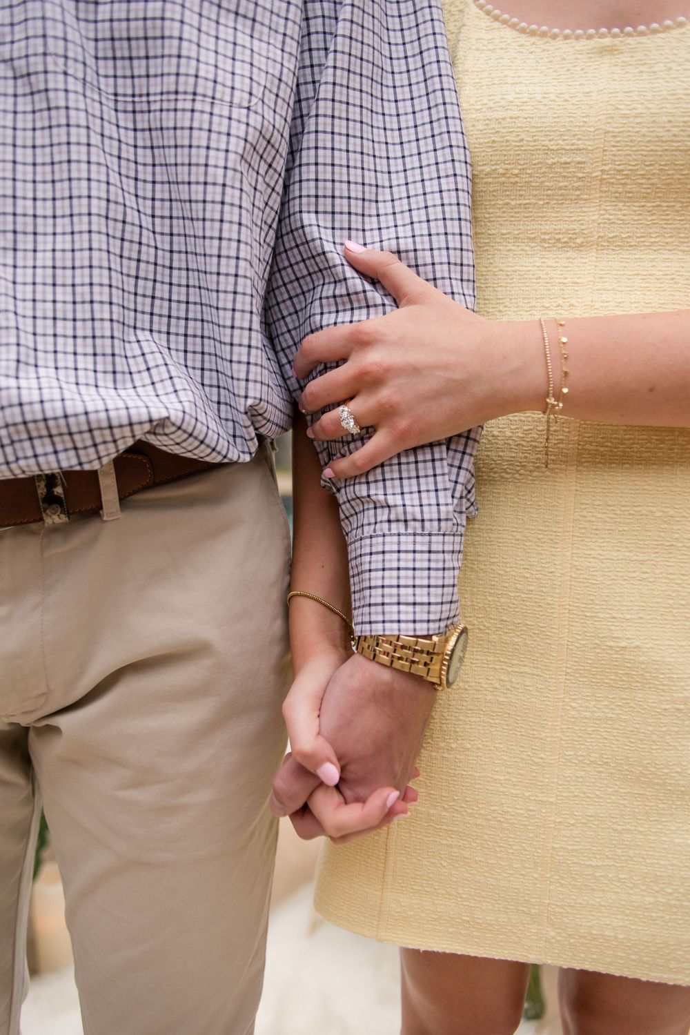 Close-up of couple's hands touching, with engagement ring visible against plaid shirt and cream dress.