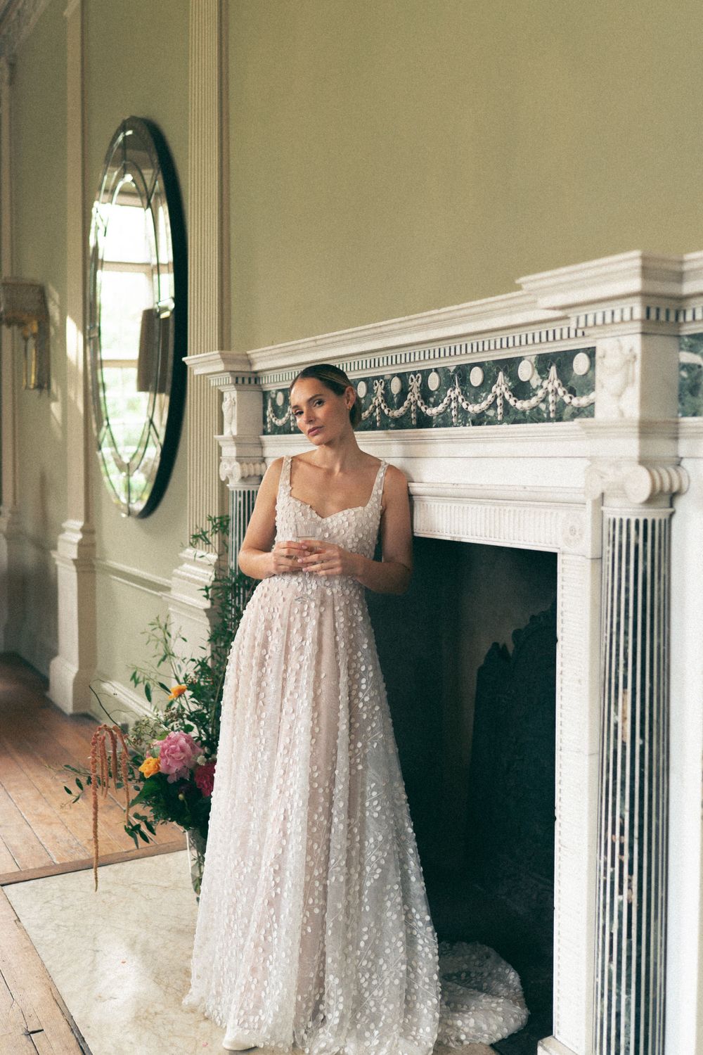 A sparkling tulle wedding gown with delicate beading stands elegantly beside an ornate white marble fireplace.