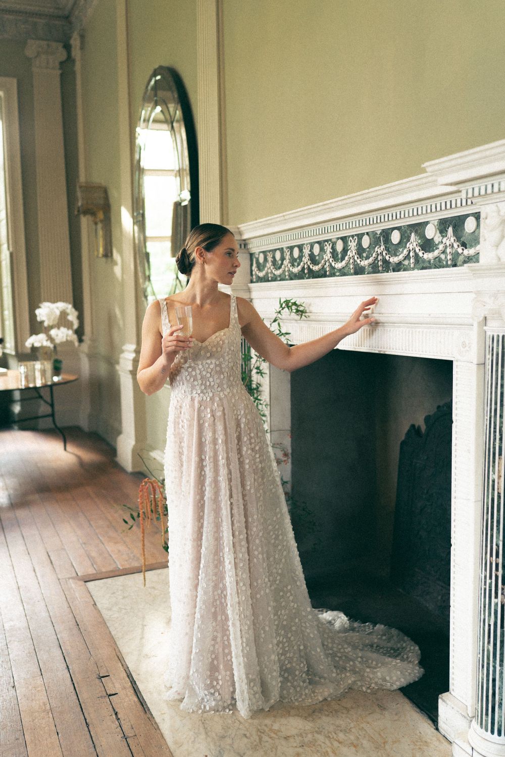 A woman in a flowing sequined wedding gown stands elegantly beside an ornate white marble fireplace in a historic interior.