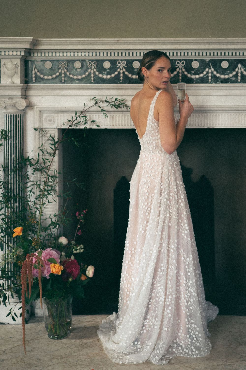 Elegant lace wedding gown with glittering details and low back design displayed in front of ornate marble fireplace with flowers.