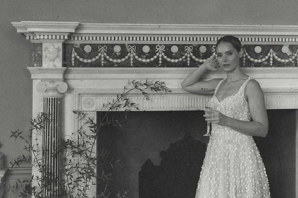 A person in a sleeveless white lace dress poses beside an ornate white fireplace mantel with decorative columns.