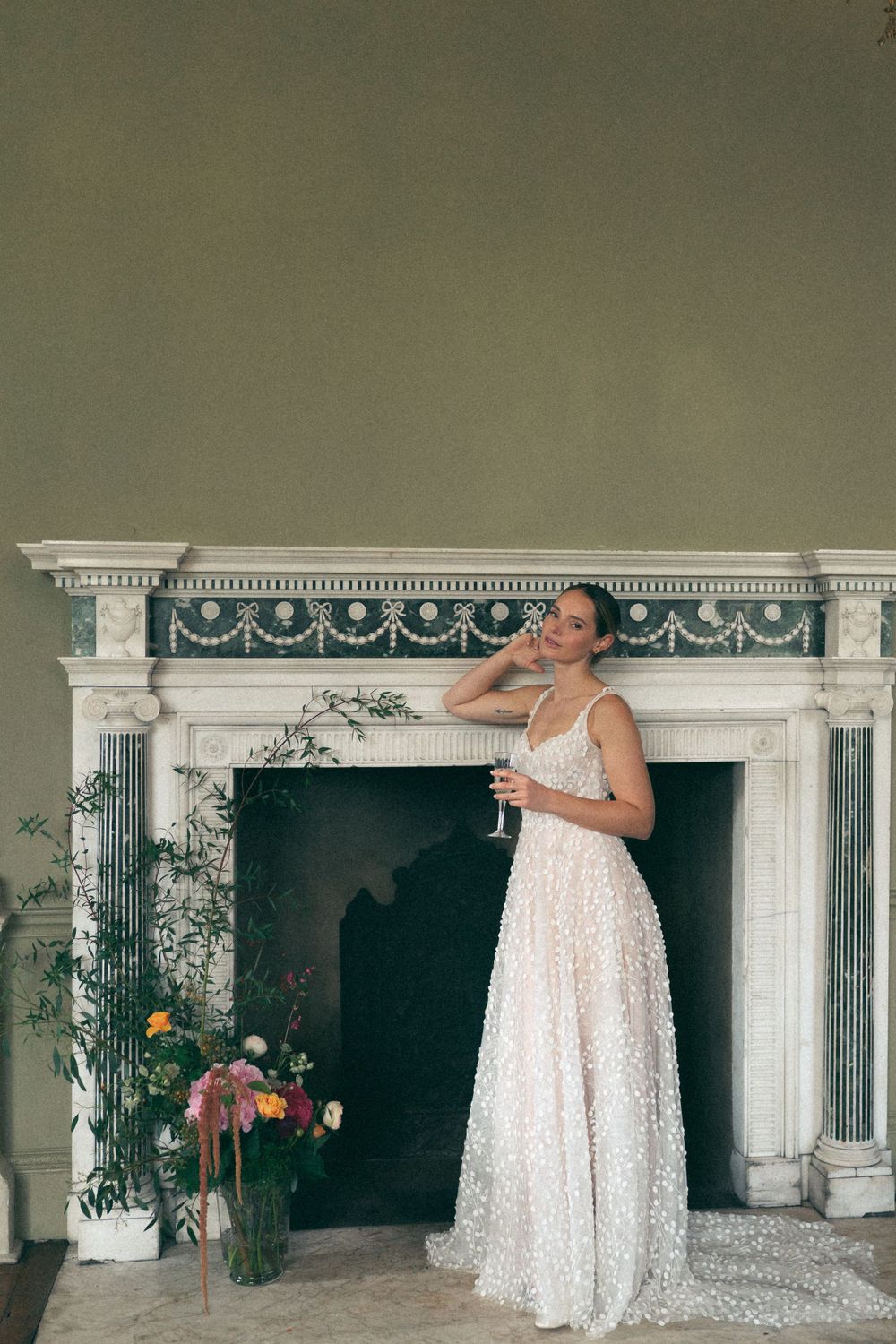 Elegant white lace dress worn in front of an ornate marble fireplace with floral decorations and classical details.