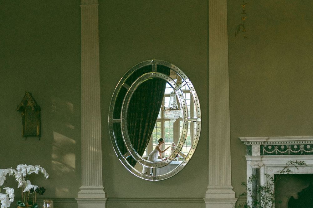 Elegant round mirror with decorative frame mounted on sage green wall beside white columns and ornate fireplace.