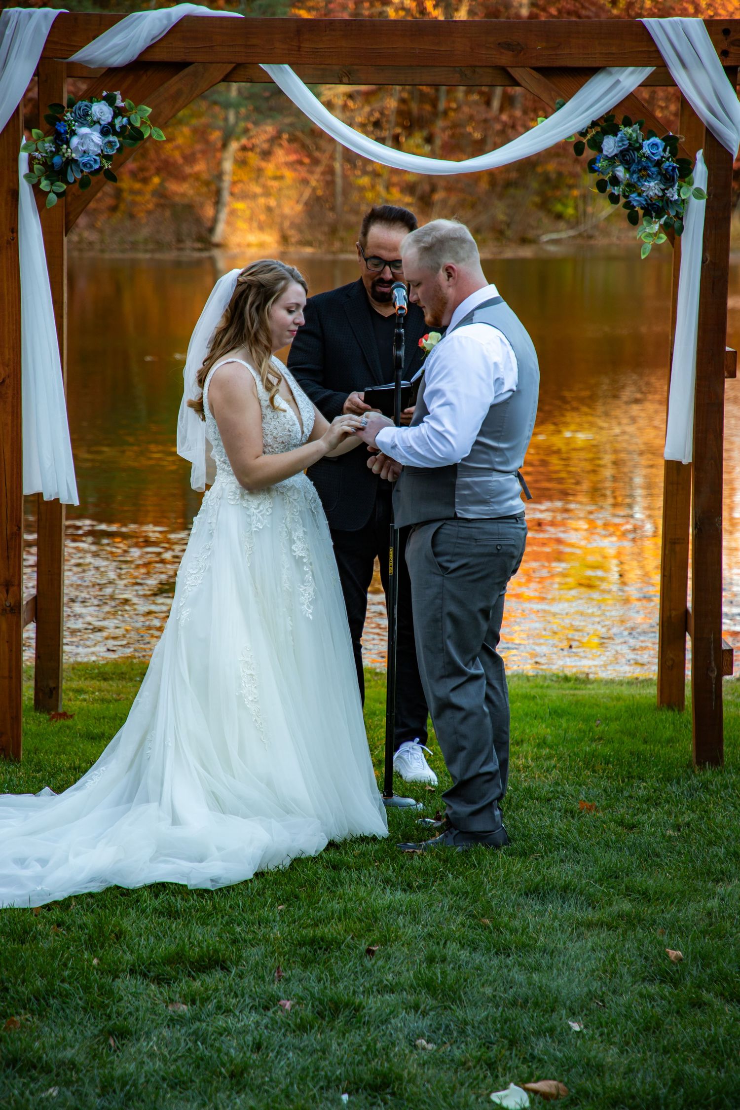 An autumn wedding ceremony takes place under a white draped arch beside a lake with golden reflections and fall foliage.