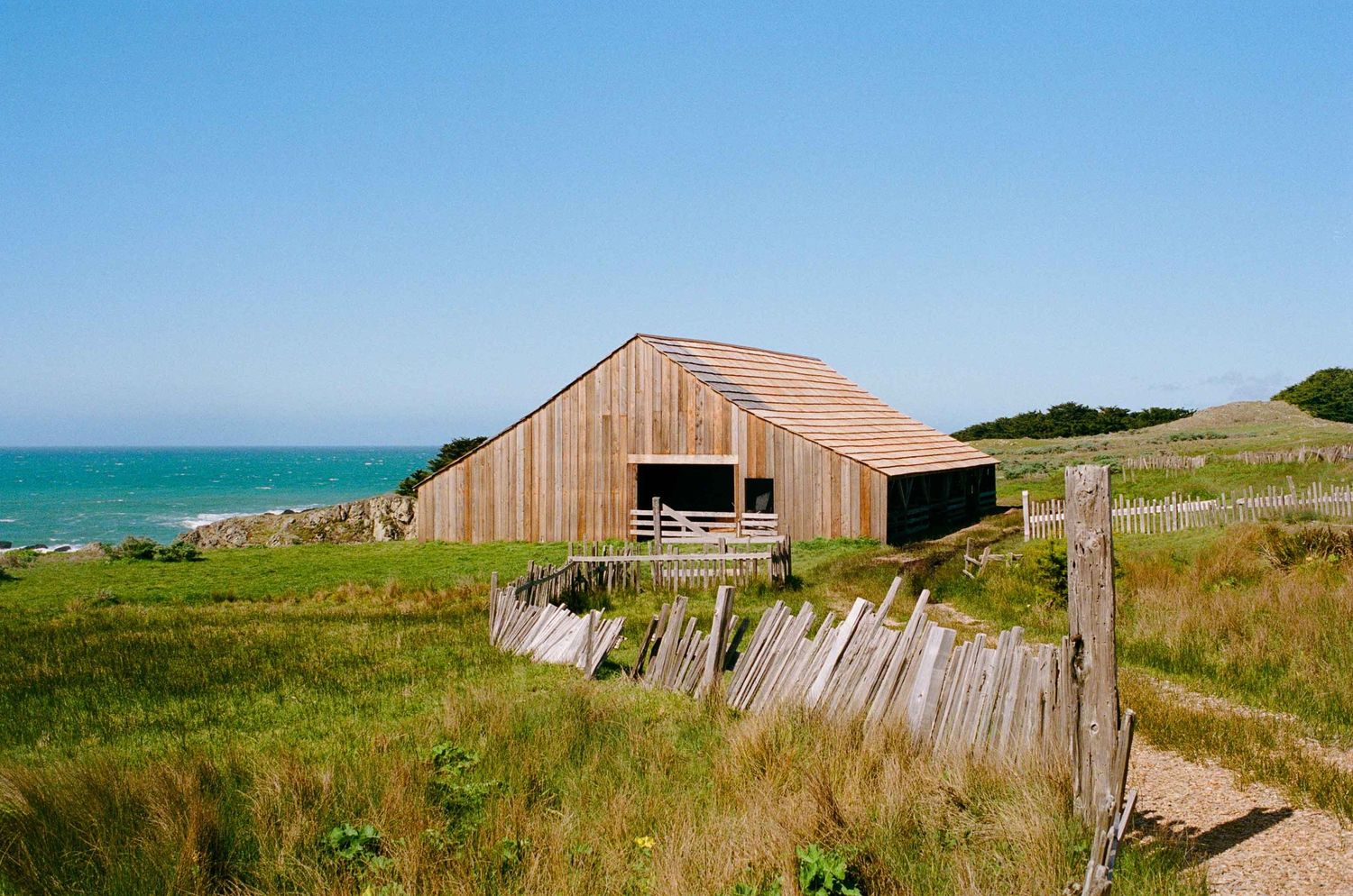 Rustic Sea Ranch wooden barn sits on grassy coastal hillside overlooking blue ocean waters on a sunny day.