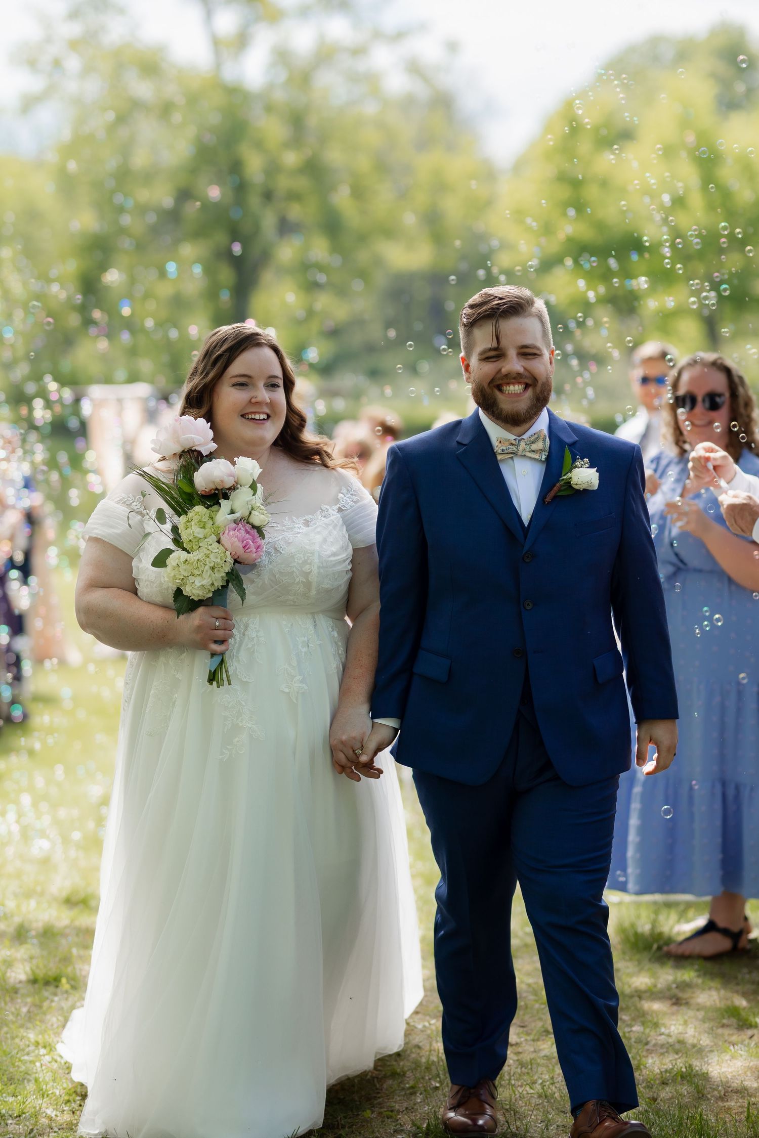 A newlywed couple walking outdoors with confetti falling around them, the bride in white holding a bouquet against a natural backdrop.