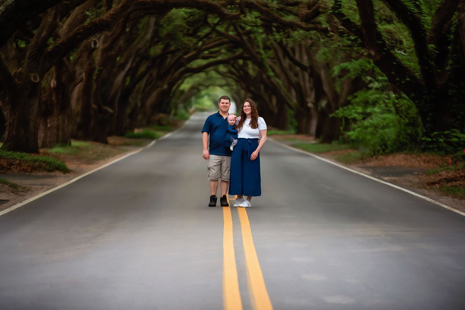 A couple stands on a tree-lined asphalt road surrounded by green forest for family portraits