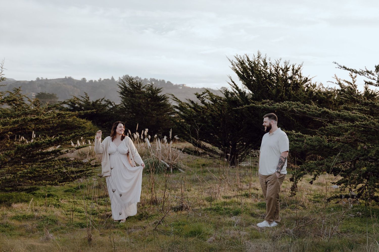 Romantic couple in white outfits sitting on blanket near tall pampas grass during outdoor photoshoot at sunset.