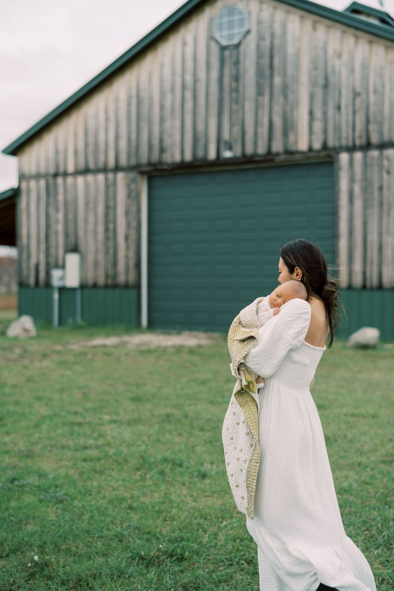 Woman in white dress holding baby stands in front of rustic barn with green door.
