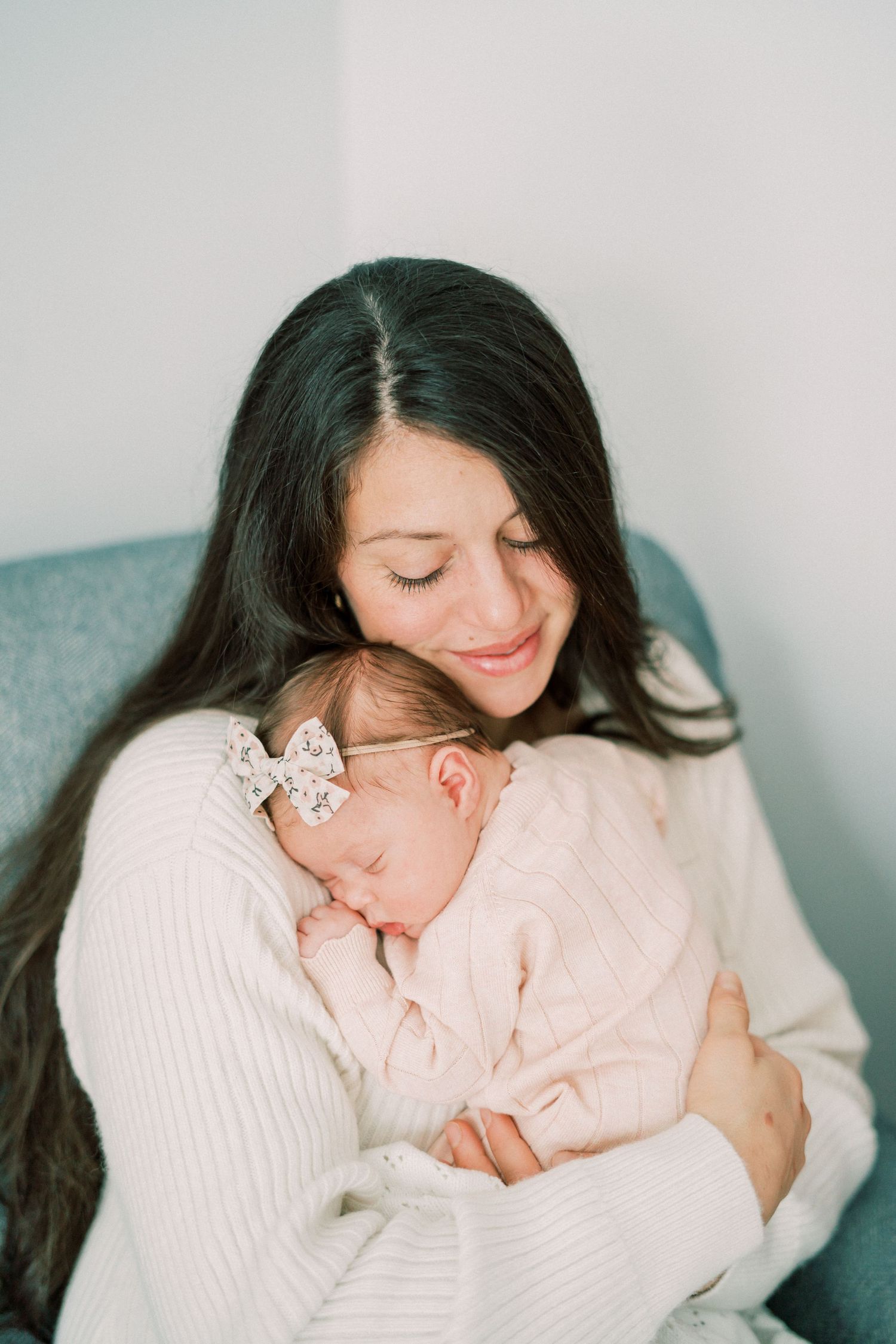 Tender moment of mother and sleeping newborn in soft white clothing by window.
