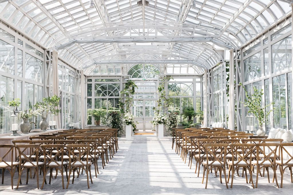 Bryllup på Norrcikens trädgårdar Båstad. Sunlit greenhouse wedding venue with wooden cross-back chairs set up for ceremony.