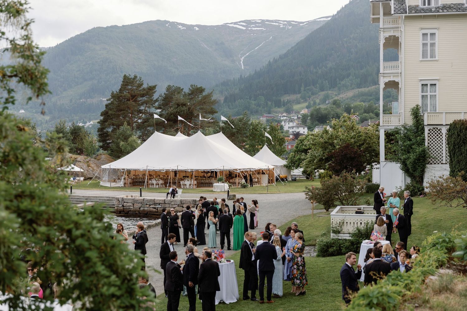 Outdoor wedding reception with white tent and mountain backdrop as guests gather on the lawn.