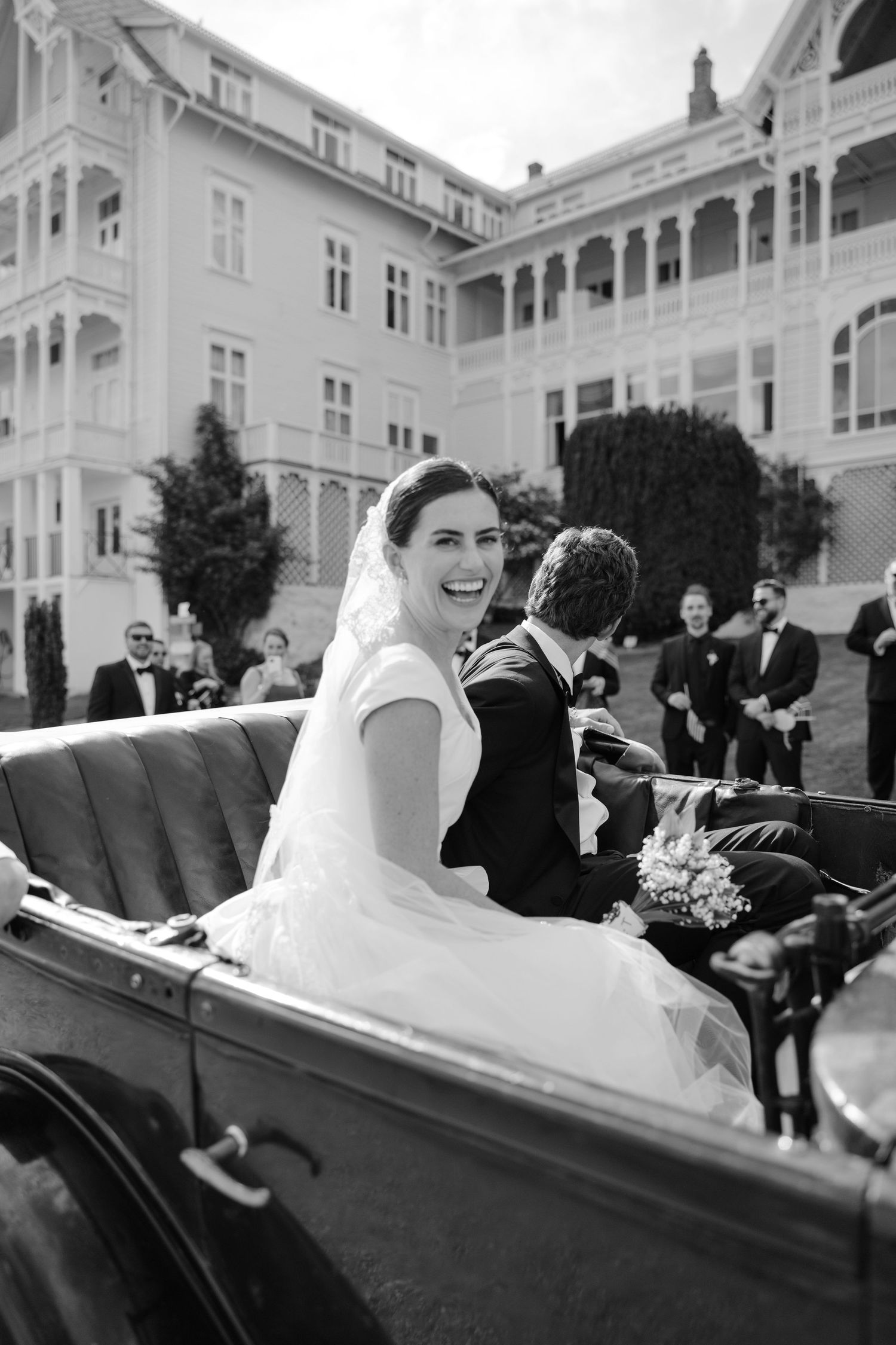 Vintage car carrying newlyweds outside an elegant historic hotel building in Norway.