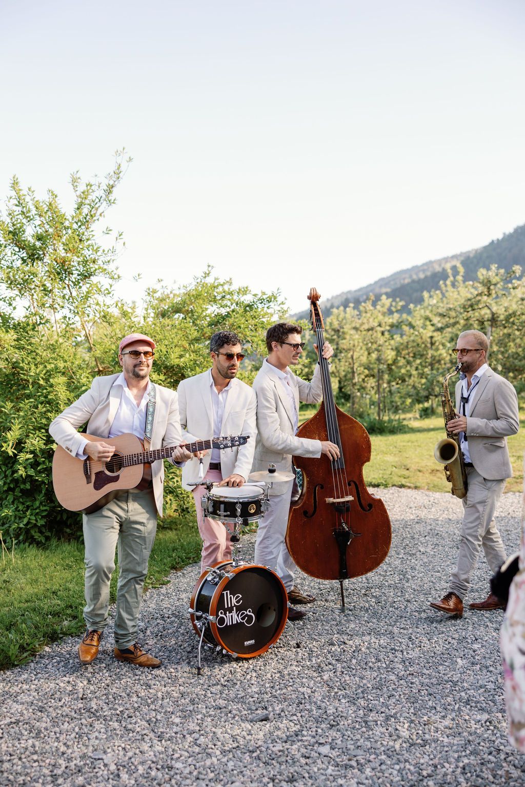 Four-piece band in light colored suits performing with instruments on a gravel path.