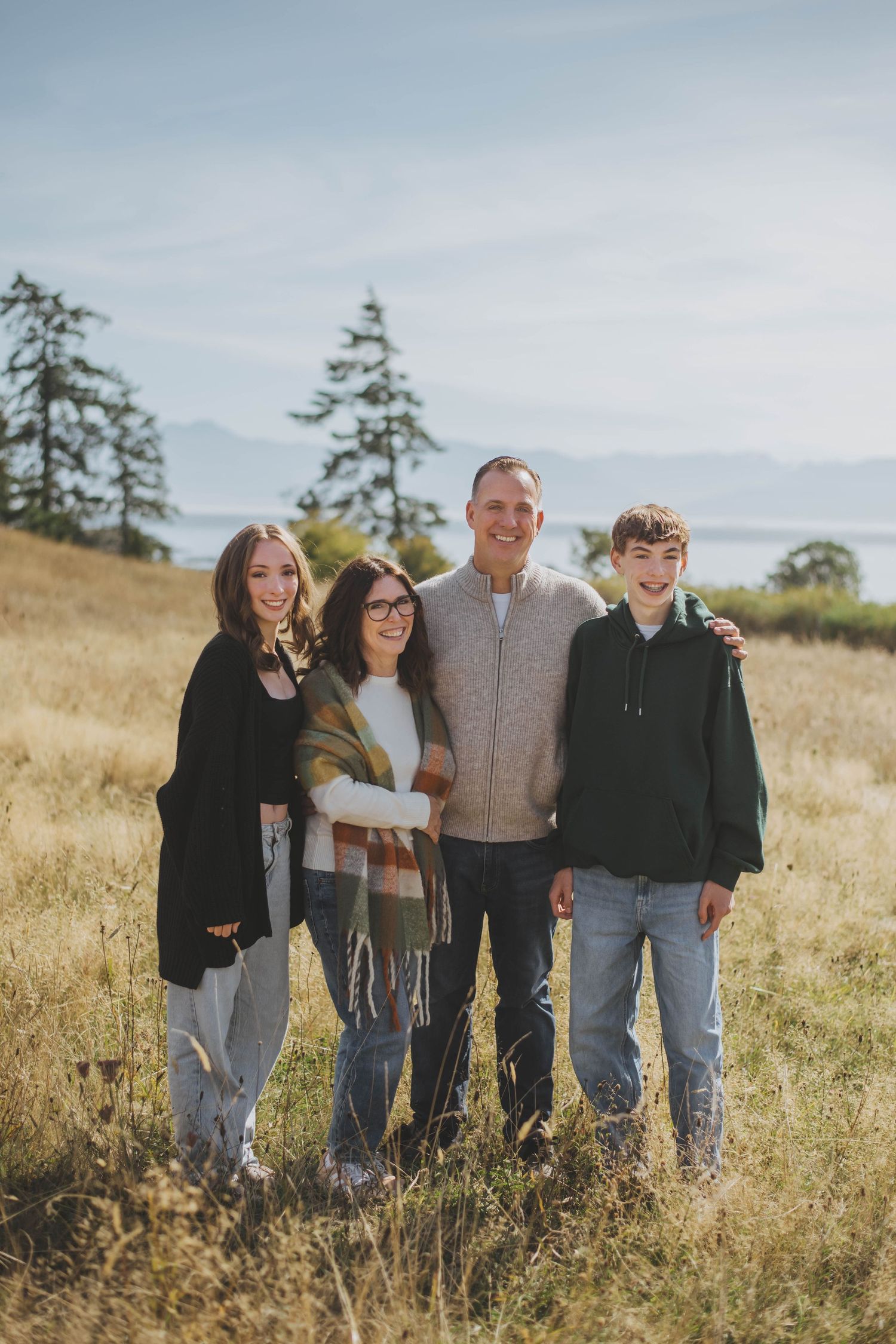 A family of four stands together in a sunlit grassy field, smiling and embracing, with trees and a distant view of mount