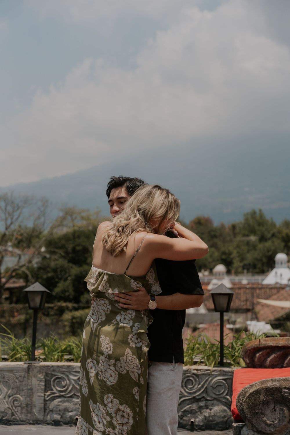 A couple embraces on a scenic terrace overlooking mountains and trees, with decorative stone walls and lanterns in view.