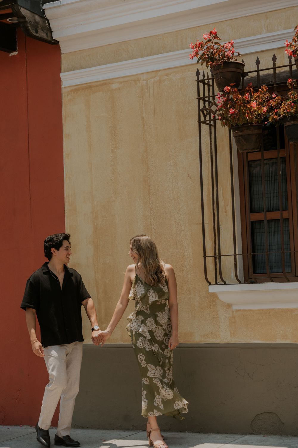 Couple holds hands while walking along colorful stucco wall with wrought iron details and hanging floral baskets.