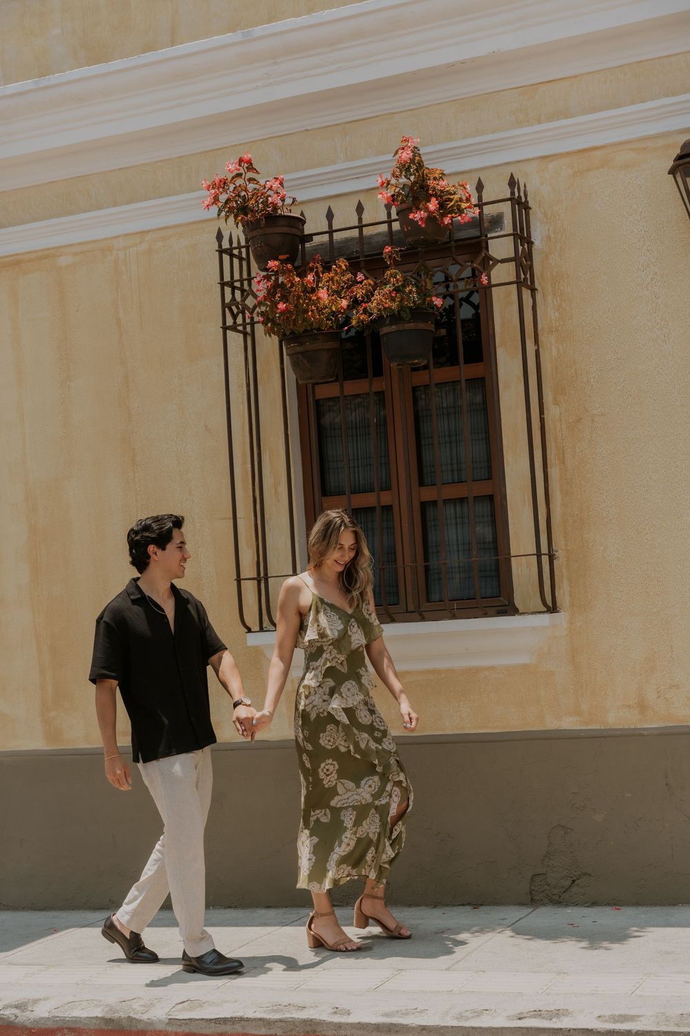 Couple walking hand in hand on a sunny day past a Spanish-style building with floral window boxes.