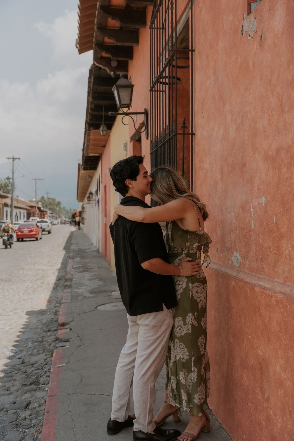 A couple embracing against a coral-colored wall on a rustic cobblestone street.
