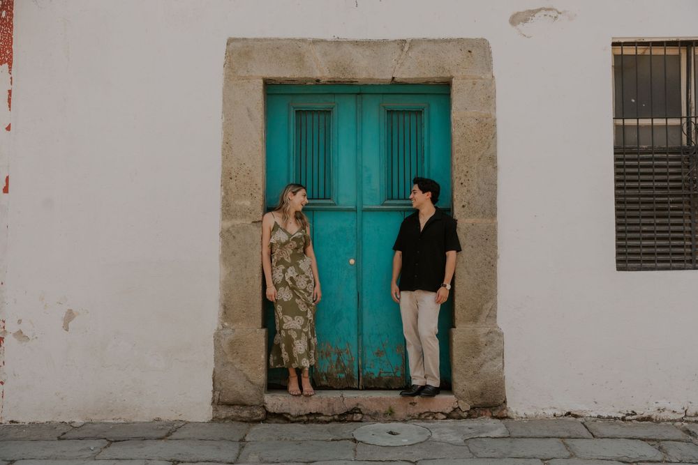 Two people stand in doorway with bright turquoise doors against white stone wall in Mexico.