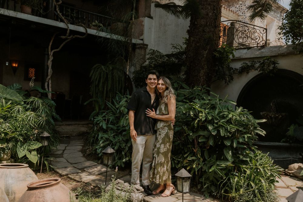 A couple embraces on a garden patio surrounded by lush tropical plants and rustic stone walls.