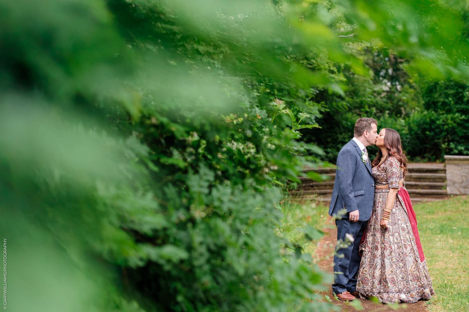 A couple in formal attire embraces in a garden setting with lush green foliage in the foreground.