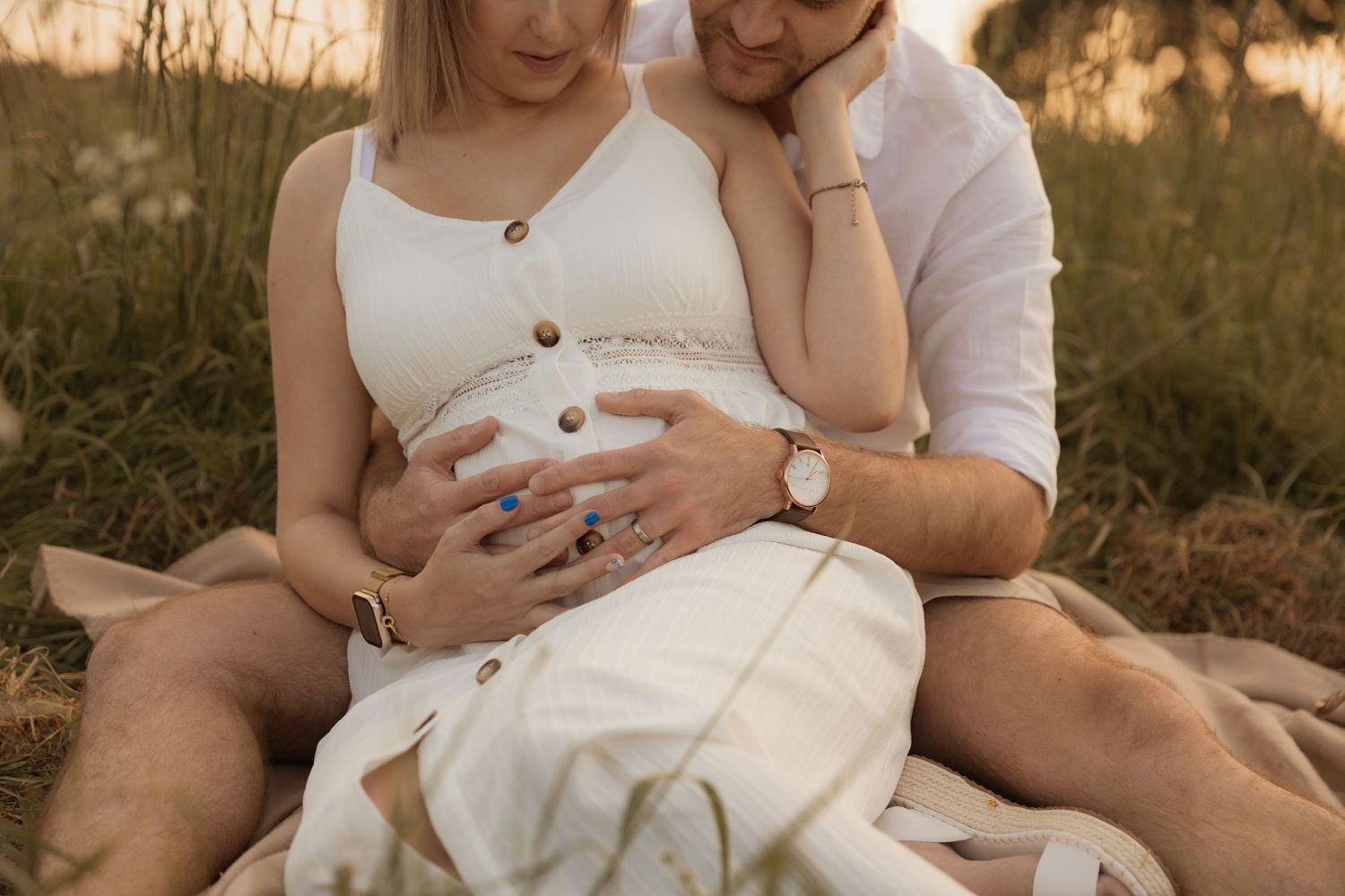 A couple sits together in a sunlit field, with gentle hands resting on a pregnant belly during maternity photoshoot.