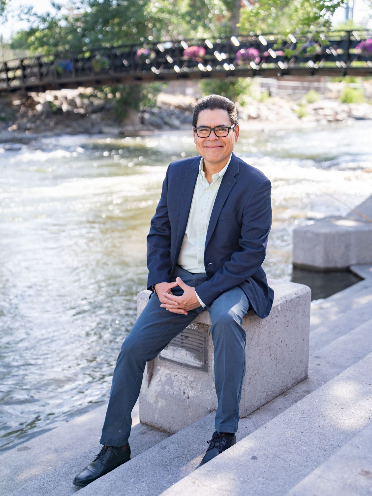 Professional wearing a navy blazer and glasses sits outdoors near water and a wooden bridge on a sunny day.