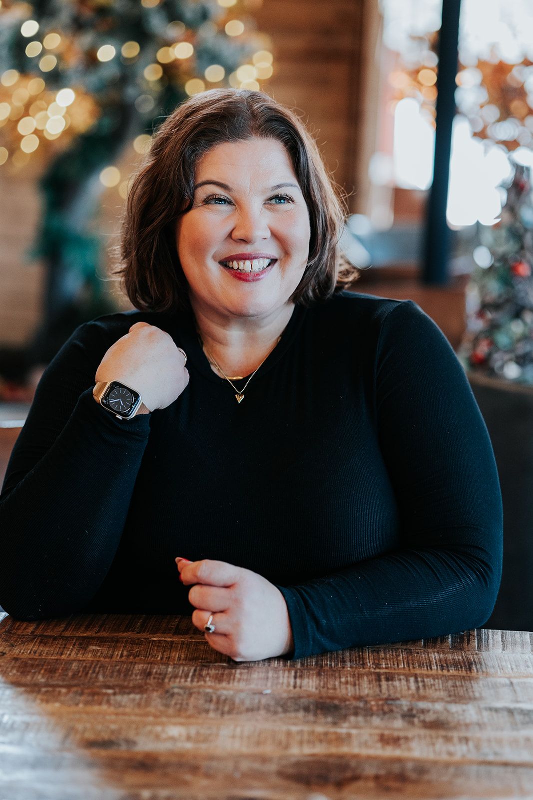 A smiling person in a black sweater sits at a wooden table with twinkling holiday lights in the background.