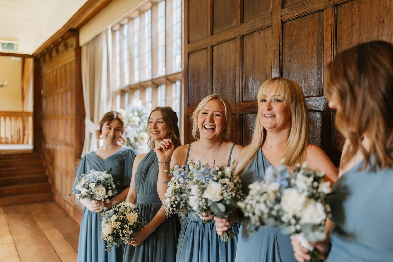 Bridesmaids in light blue dresses share joyful laughter while holding white and green floral bouquets in a wood-paneled room.