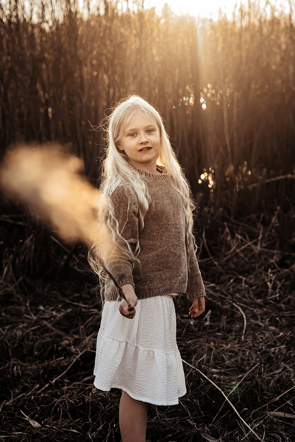 Porträtt på en ung flicka med långt blondt hår som håller ett vasstrå i sin hand som motljuset silar sig igenom, fotograf Lets Bloom Media.