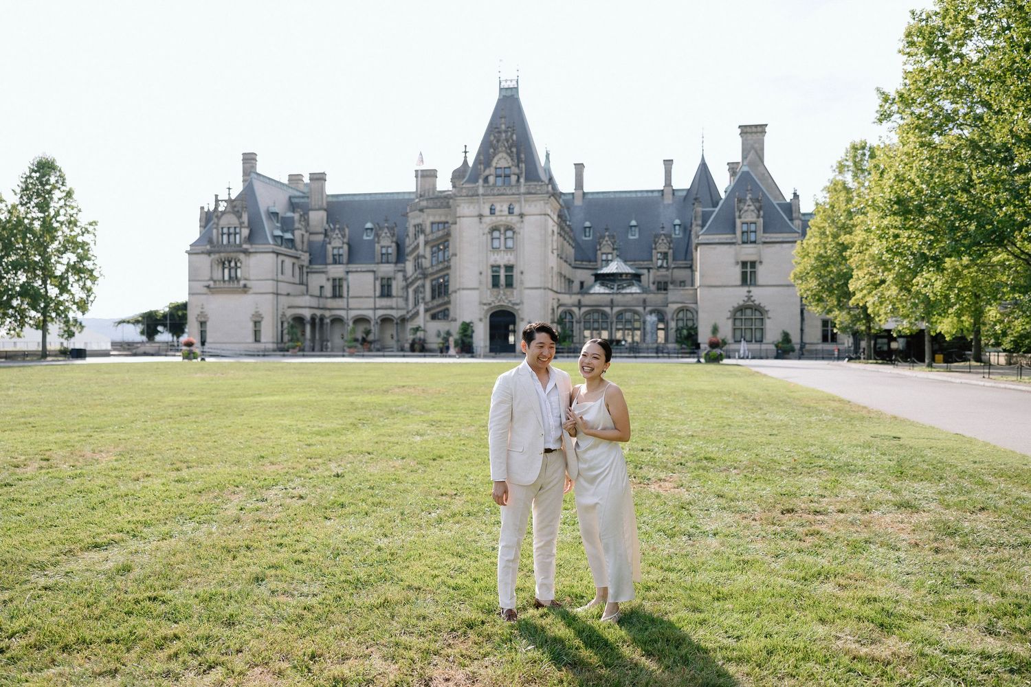 A couple stands on a manicured lawn in front of the grand Biltmore Estate mansion on a sunny day.