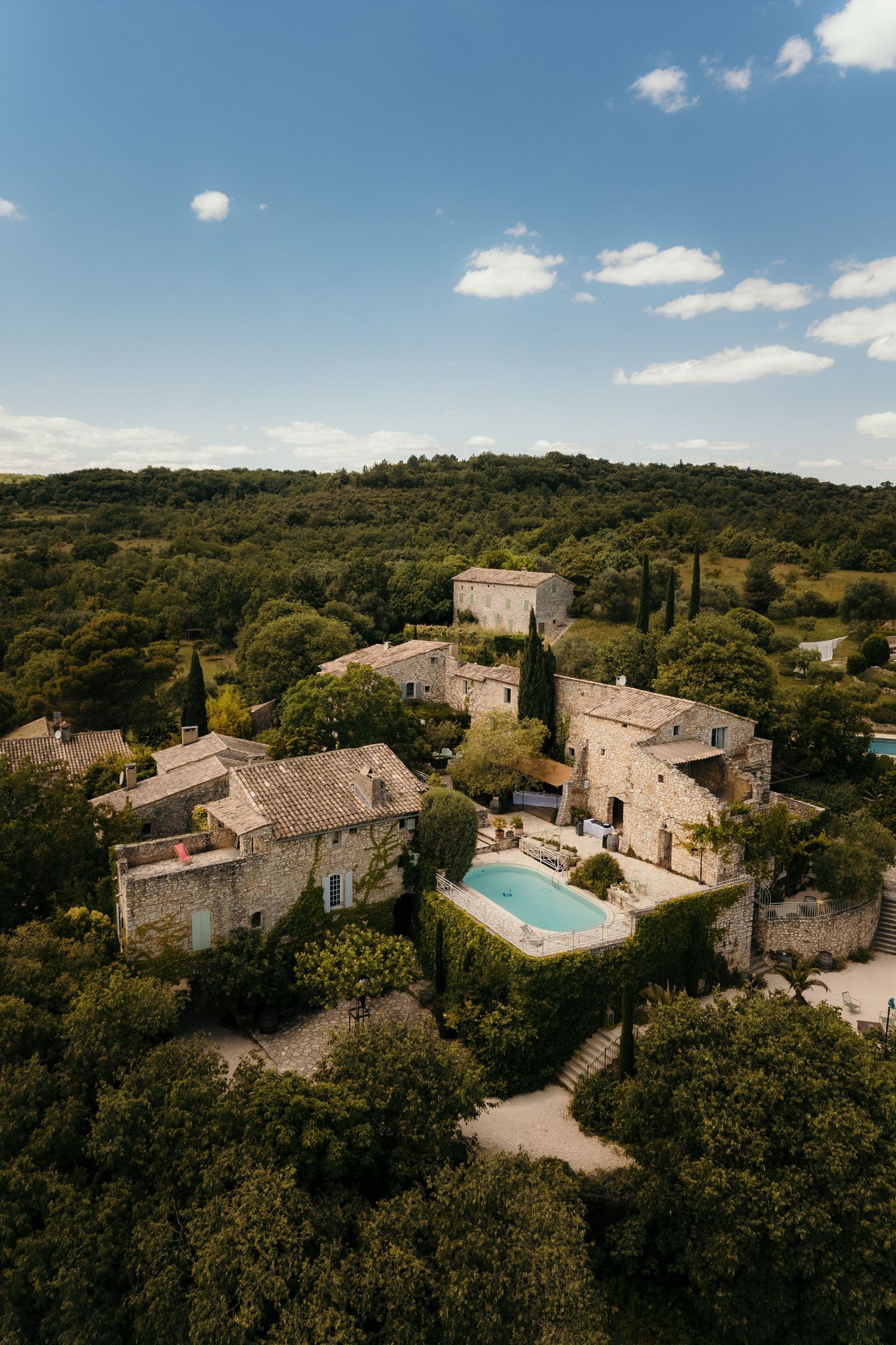 Aerial view of rustic stone villa with swimming pool nestled in lush Mediterranean countryside under blue sky.