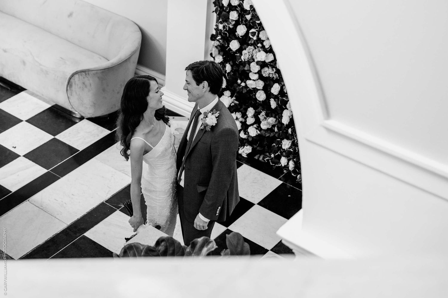 A couple stands on a black and white checkered floor near a curved wall with floral decorations in monochrome style.