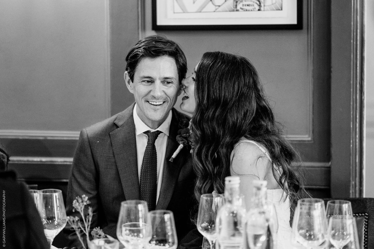 A couple shares a candid black and white moment at an elegant dinner reception with wine glasses on the table.