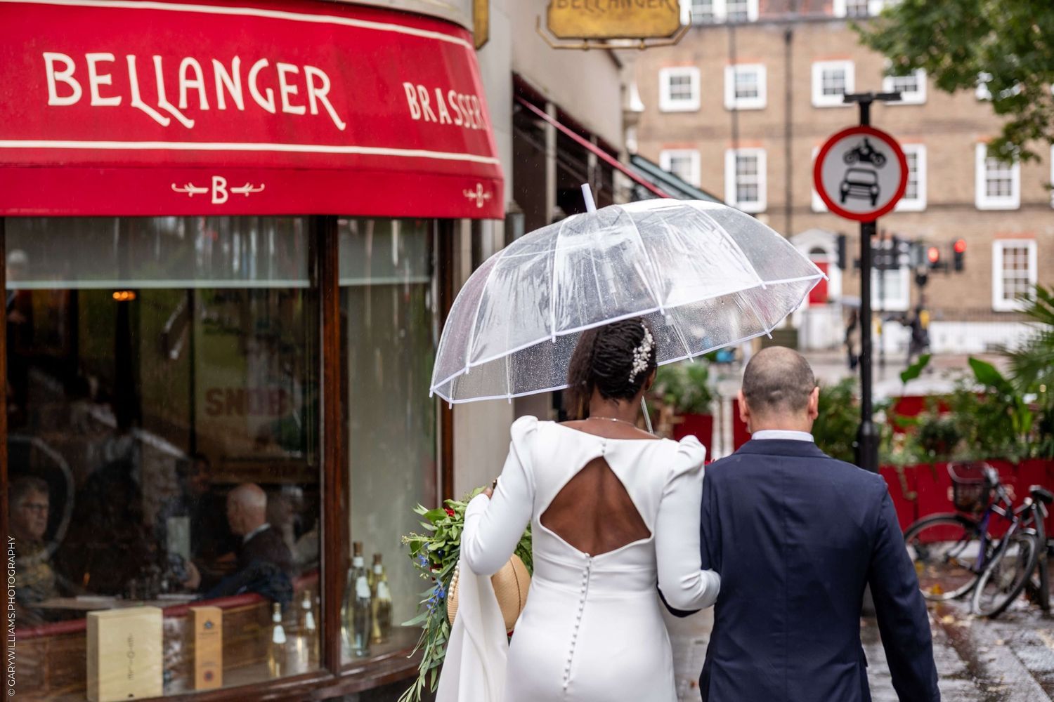 A person with a clear umbrella walks past Bellanger restaurant with its red awning on a city street.