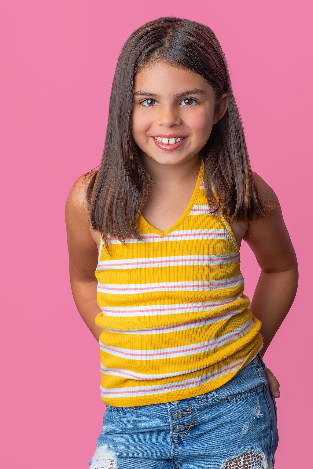 Young person in yellow and white striped tank top and denim shorts poses against pink background.
