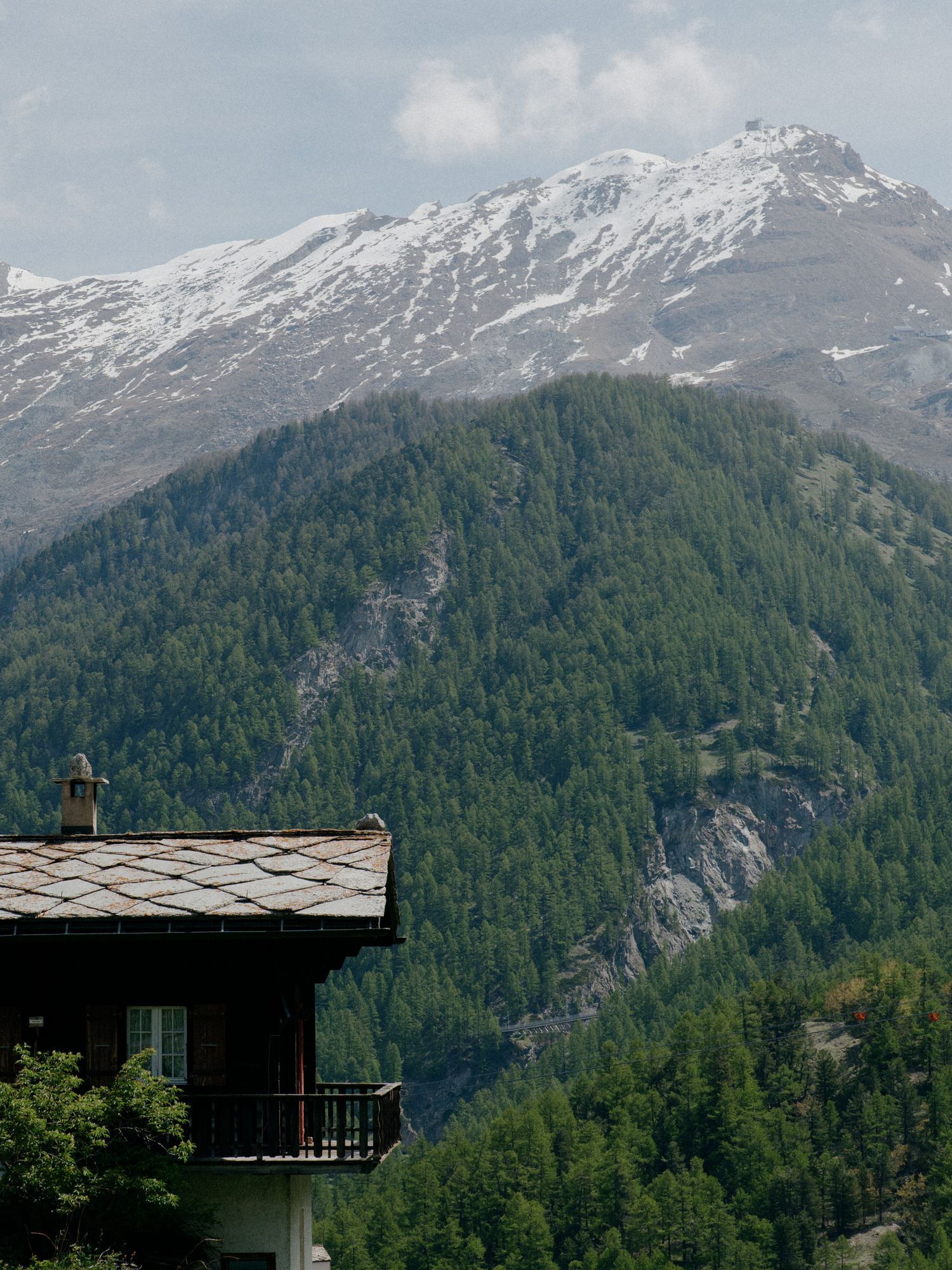 A rustic mountain chalet with a snow-capped Alpine peak and dense evergreen forest in the background.