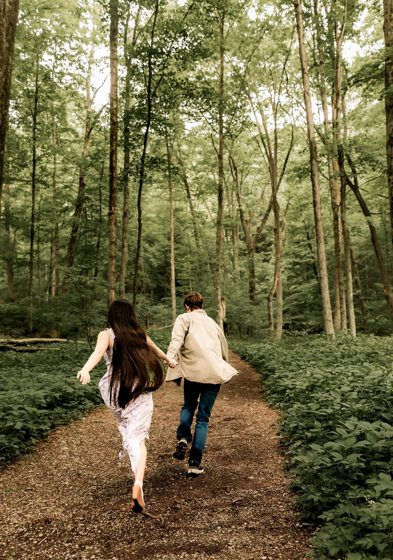 Two people joyfully run together on a winding forest path surrounded by lush green trees and foliage.