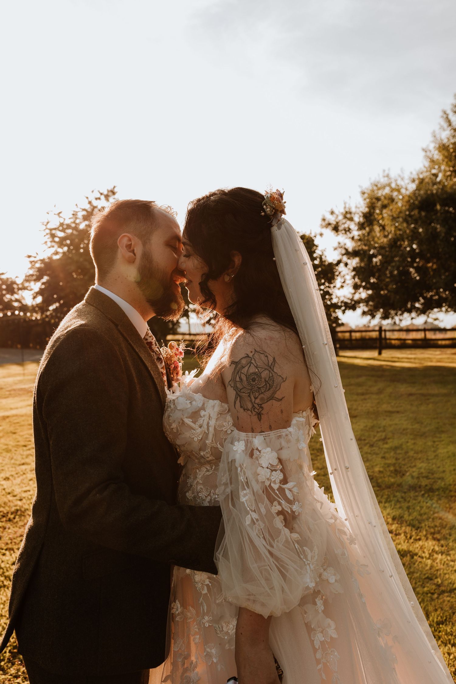 Newlywed couple shares an intimate moment at sunset in a golden-lit field with a flowing white lace dress and veil.