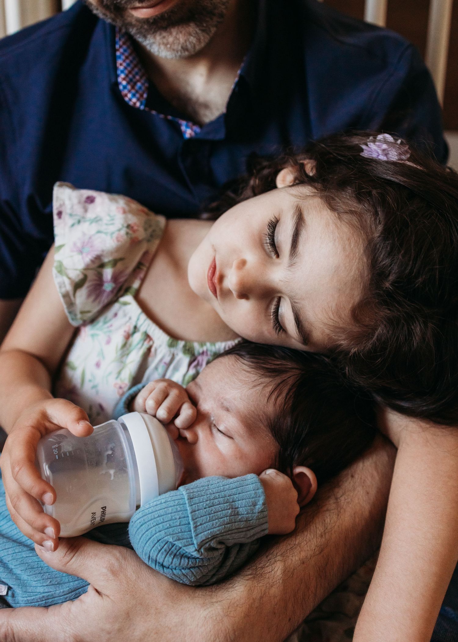 Young child in floral dress rests peacefully while holding baby's bottle as infant drinks in relaxed pose.